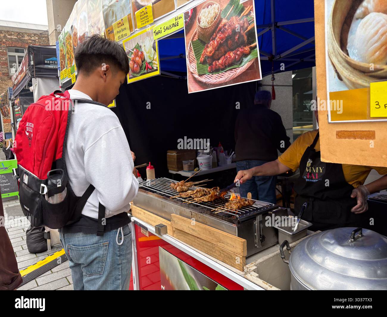 Mercato del sabato Britomart, Auckland, nuova Zelanda. Un venditore di Street food griglierà gli spiedini mentre un cliente guarda. I segni per la salsiccia tailandese sono visibili, ciao Foto Stock