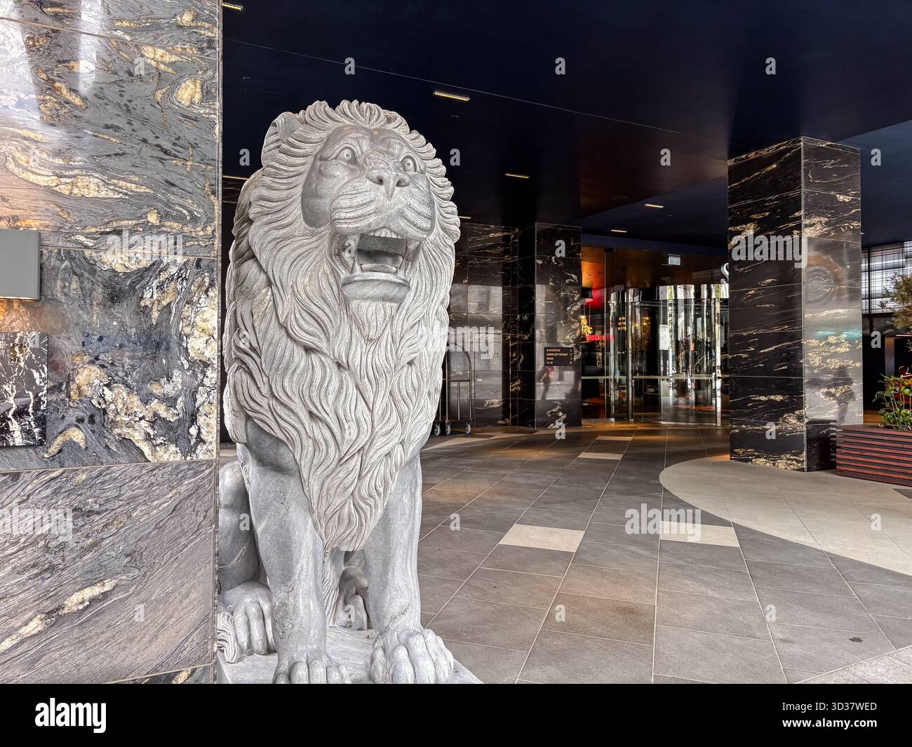 Statua del leone di pietra a bocca aperta sorge in una lussuosa lobby moderna, circondata da colonne di marmo scuro lucido e da elegante pavimento piastrellato ad Auck Foto Stock