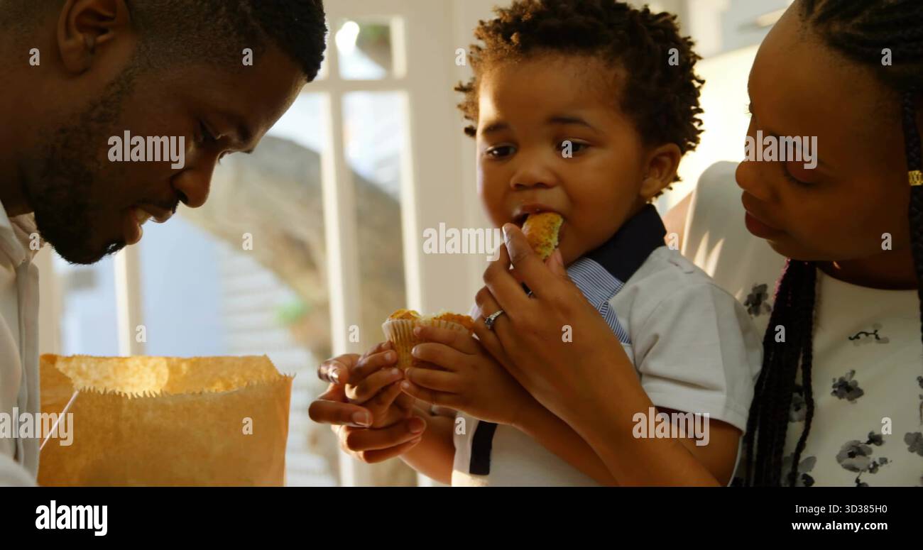 I genitori che danno da mangiare ai muffin dei bambini in cucina accanto a una grande finestra che mostra il tronco dell'albero, con sacchetto di carta Foto Stock
