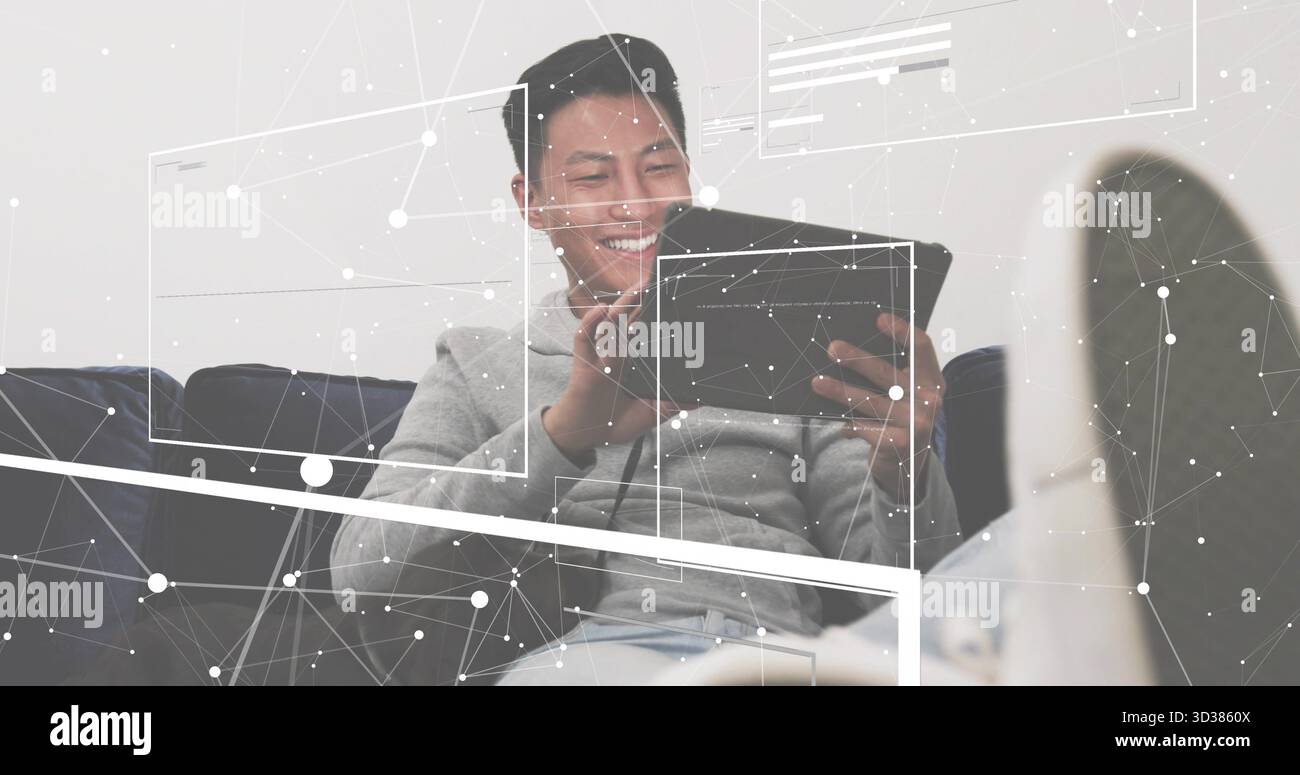 Uomo sorridente in felpa con cappuccio che tiene il tablet appoggiato sul divano scuro di casa, con grafica di rete Foto Stock