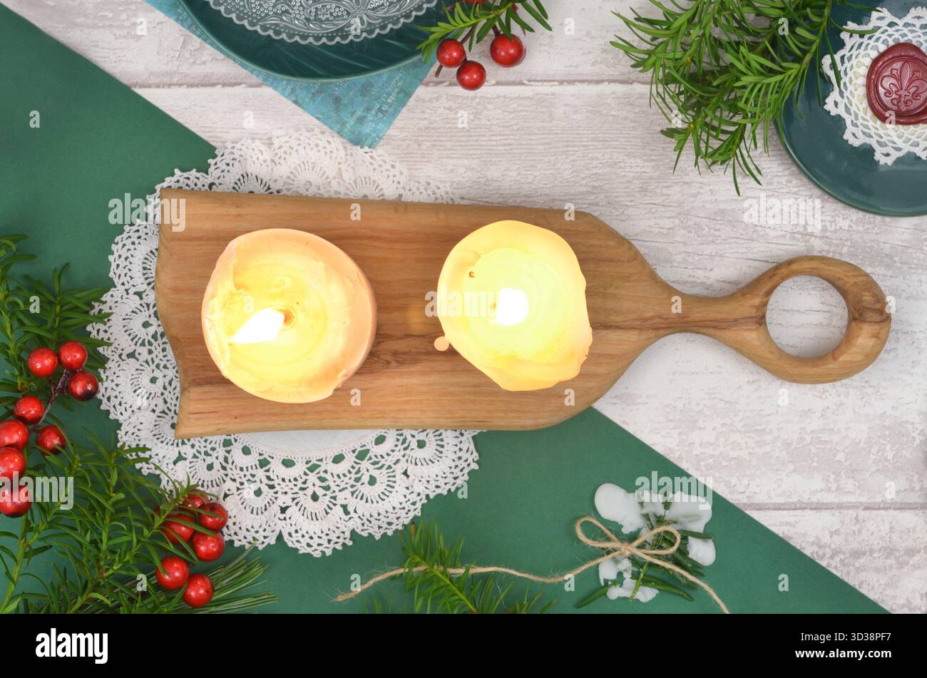 Due candele accese su tavola di legno, decorazioni natalizie, tovaglioli all'uncinetto, francobolli, cera, piatti, rami di conifere e bacche rosse. Foto Stock