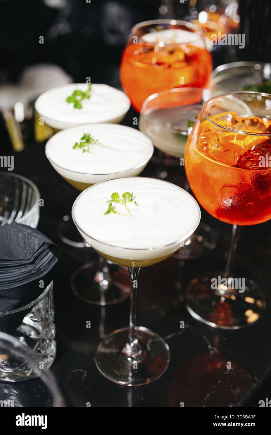 Cibo, eleganti cocktail artigianali con cime spumeggianti e decorazioni alle erbe, oltre a bevande spritz Aperol luminose su un bancone da bar scuro Foto Stock