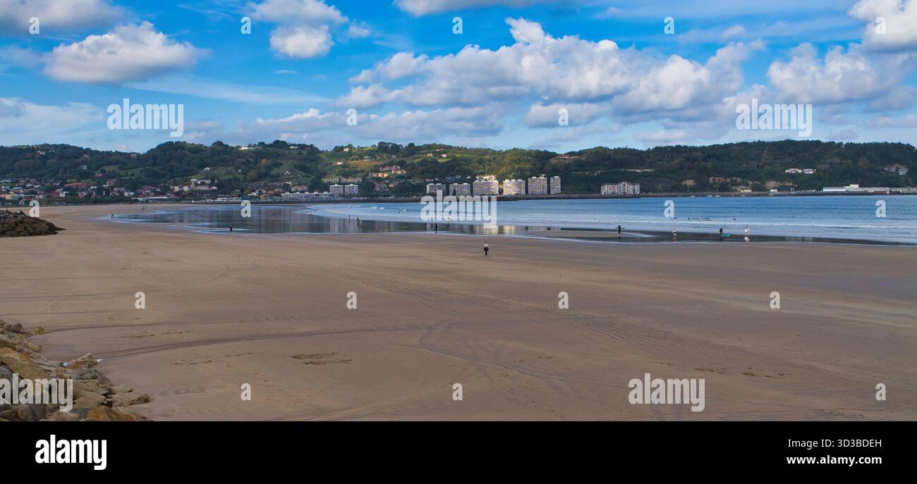 Ampie sabbie a bassa marea di Hendaye, in Francia, con la costa degli Spains sotto i cieli luminosi e le nuvole ondulate. Gli escursionisti aggiungono un tocco di grandezza e un'atmosfera tranquilla e ariosa. Foto Stock