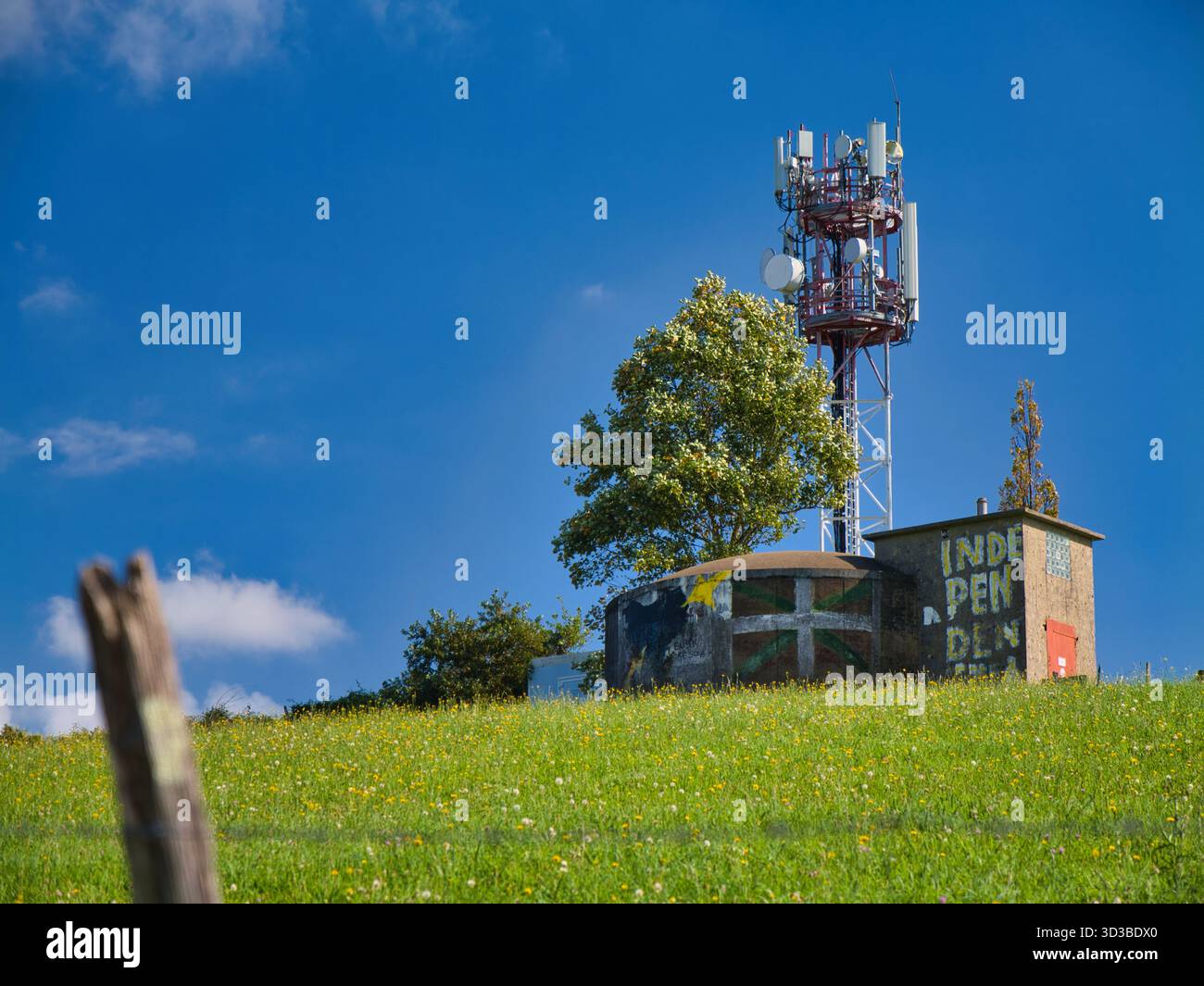Espelette, Francia - 10 ottobre 2025: A Espelette, Francia, una torre di celle in cima a una collina si affaccia su un prato e un piccolo edificio dipinto con un messaggio di indipendenza basca contro un cielo azzurro. Foto Stock