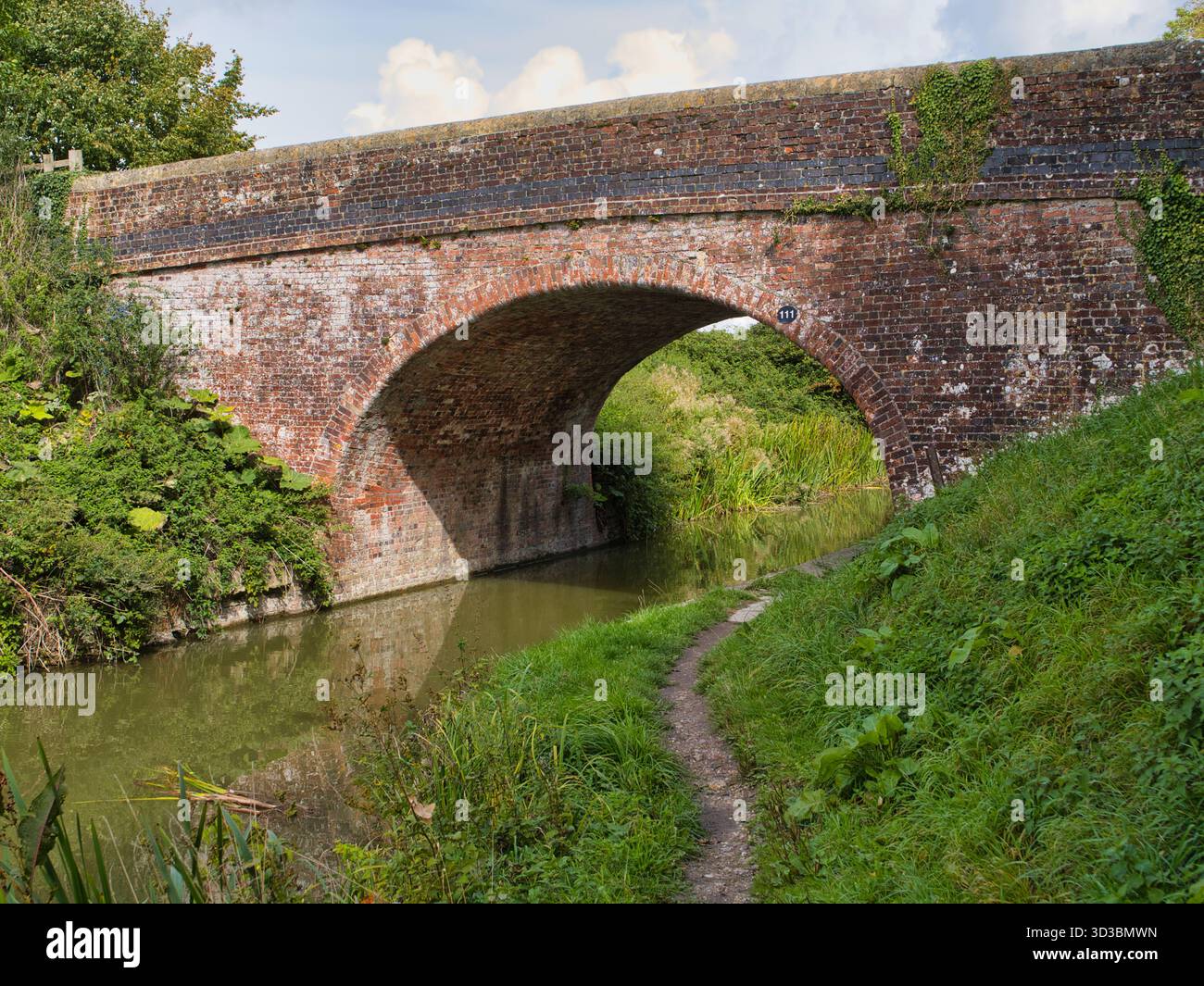 Wiltshire, Regno Unito - 09 settembre 2025: Storico ponte 111 ad arco in mattoni che si estende sul canale Kennet & Avon nel Wiltshire, Regno Unito, con un sentiero e una lussureggiante vegetazione estiva sotto la luce soffusa del pomeriggio. Foto Stock