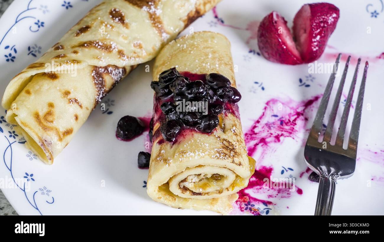 Un piatto presenta due appetitose crepes ripiene di ingredienti dolci e ricoperte di spalmazione di mirtilli selvatici. Foto Stock