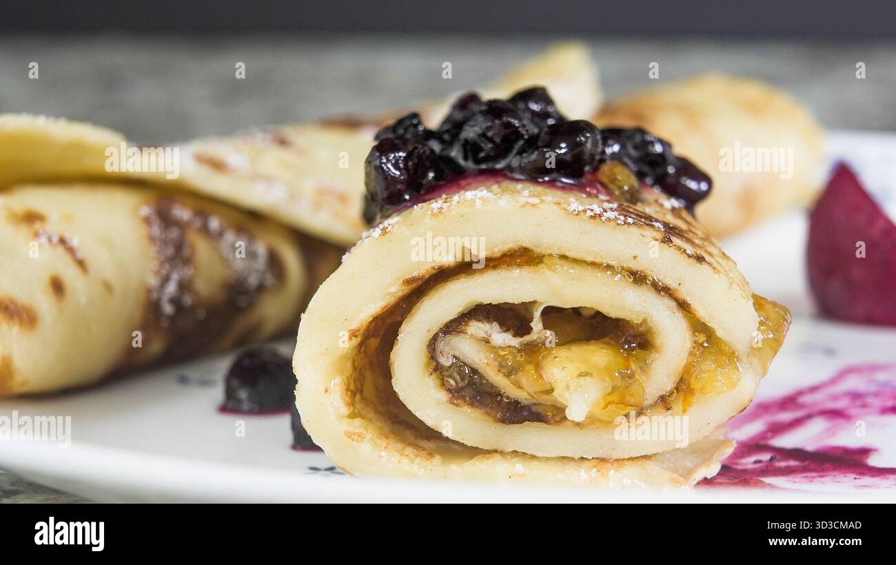 Un piatto presenta crepes arrotolate piene di marmellata di arance. Le crepes sono guarnite con un ricco spalmabile di mirtilli selvatici e servite insieme a VIBRANT Foto Stock