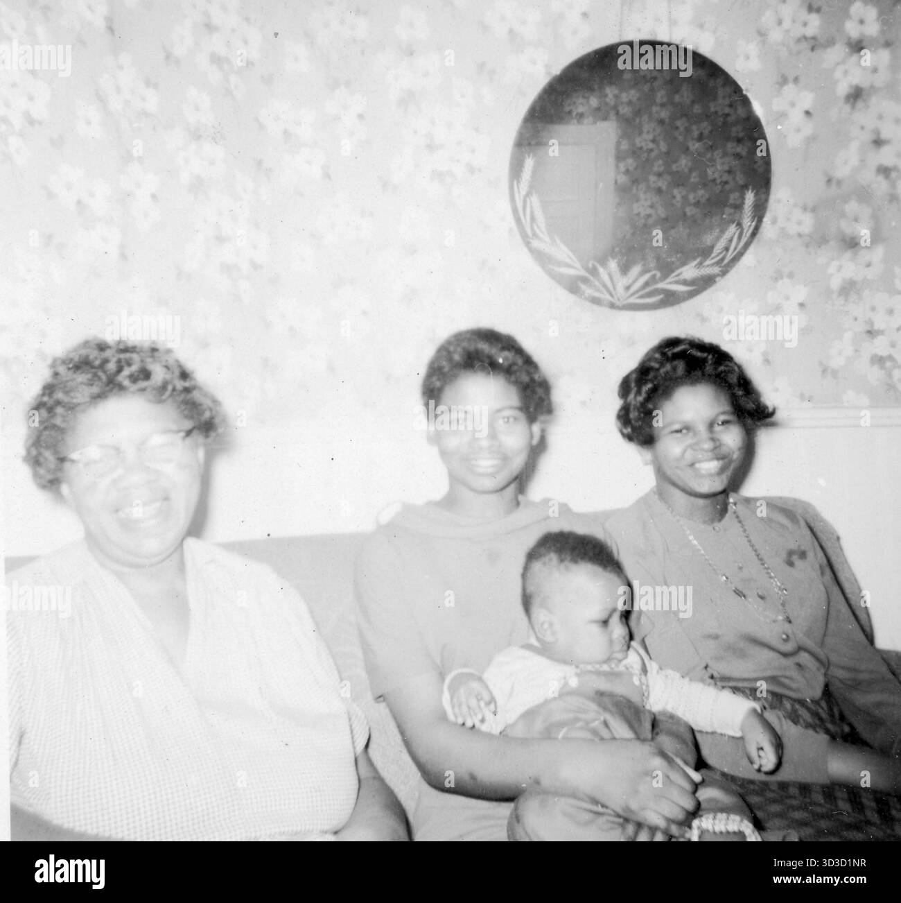 Tre donne afroamericane, probabilmente di più generazioni, si siedono vicine su un divano da salotto come una può ospitare un neonato, con carta da parati floreale e uno specchio rotondo visibile dietro di loro, Stati Uniti, 1958. Foto Stock