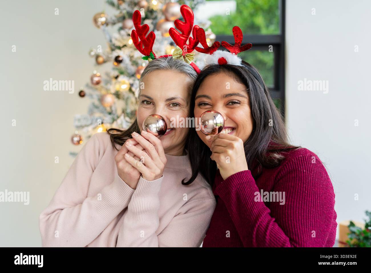 Madre e figlia diverse che tengono baule vicino al naso indossando fasce di corna accanto all'albero di Natale Foto Stock