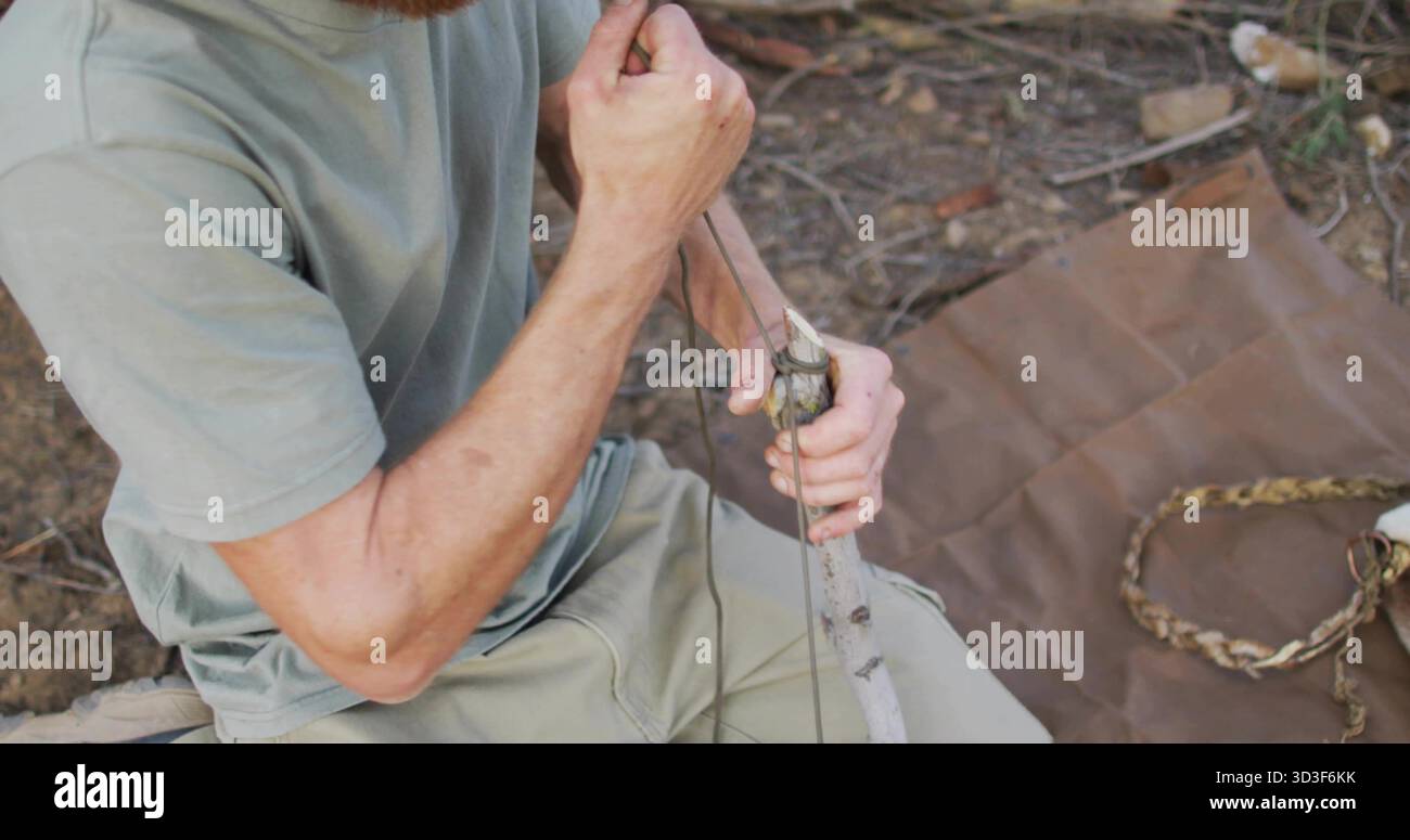 Uomo con t-shirt verde che lega la corda intorno a un palo di legno sul pavimento della foresta, con bastoncini sparsi Foto Stock