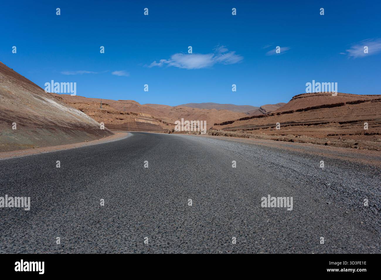 Ripresa da un angolo basso di una strada che attraversa il deserto marocchino sotto un cielo blu vivace con nuvole bianche sparse Foto Stock