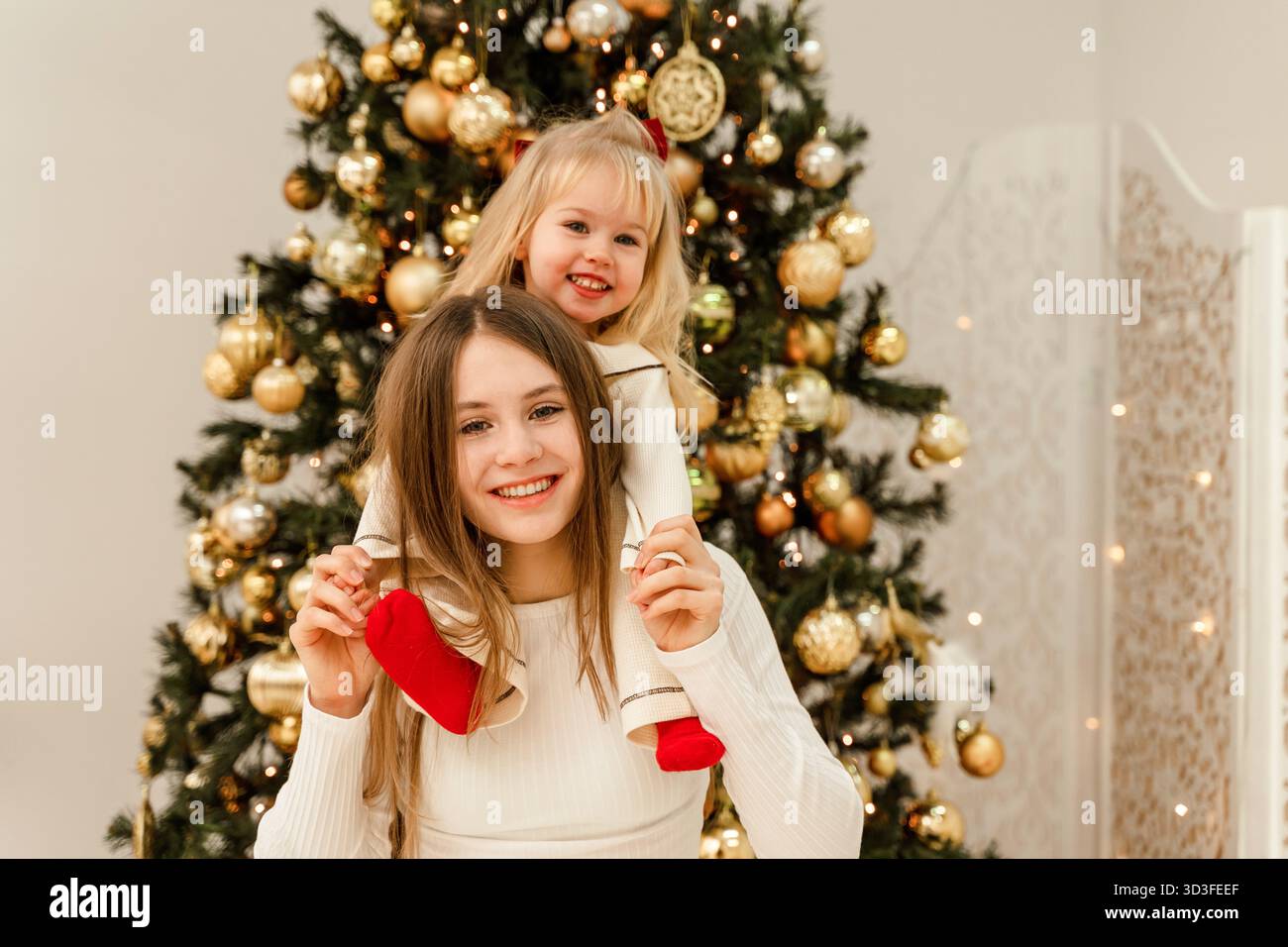 Due sorelle, una adolescente e una bambina di 3 anni, si abbracciano accanto a un albero di Natale decorato. Decorazioni per la casa per il nuovo anno. Tradizioni di famiglia Foto Stock