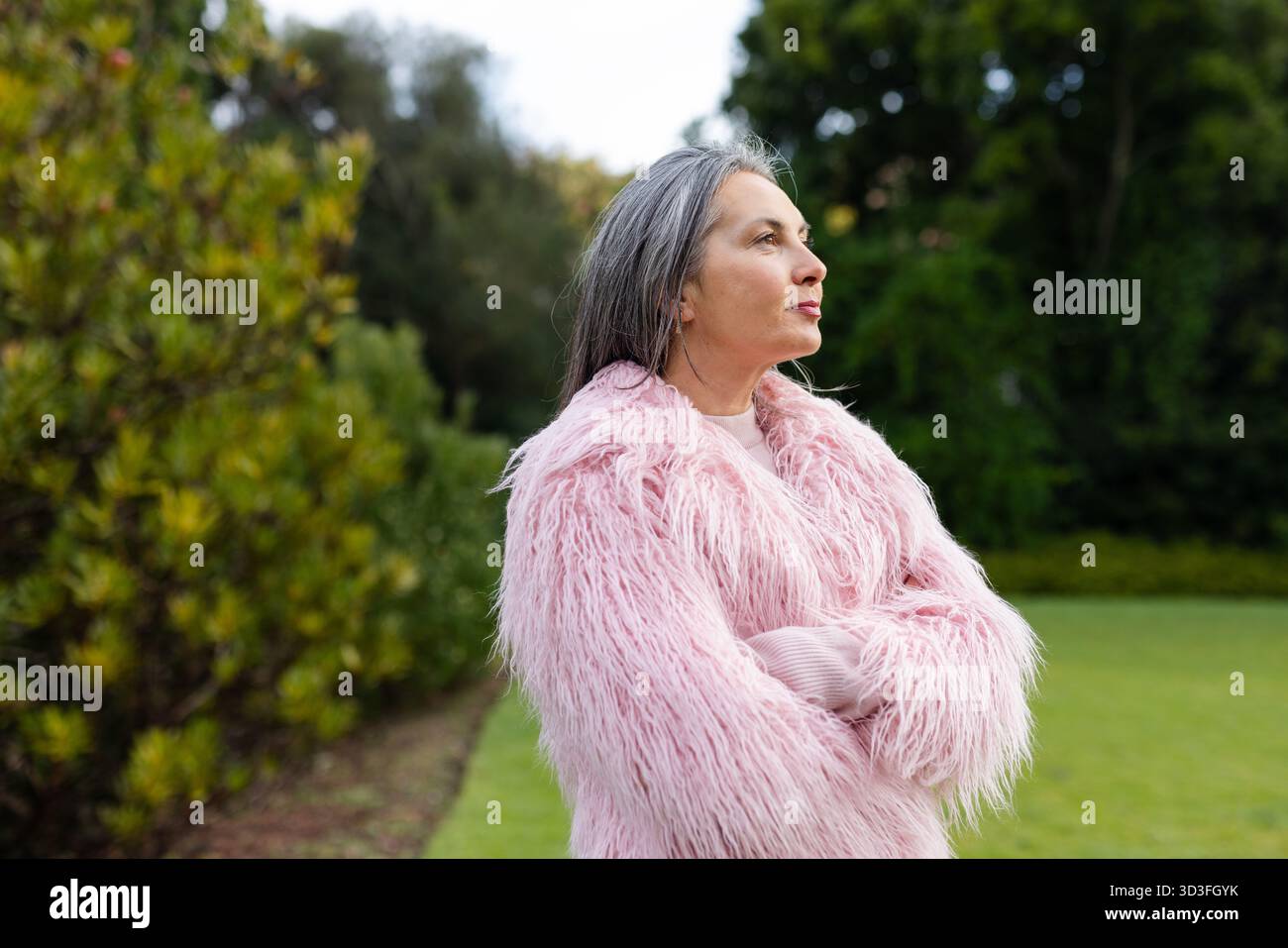 Donna anziana in piedi con le braccia incrociate che guardano direttamente in giardino indossando un cappotto di pelliccia sintetica rosa, spazio copia Foto Stock