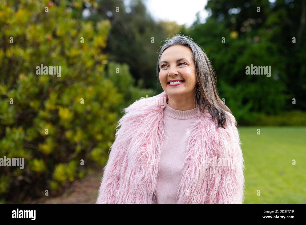 Donna che indossa un cappotto di pelliccia sintetica rosa e sorride mentre guarda a sinistra in un giardino ben curato Foto Stock