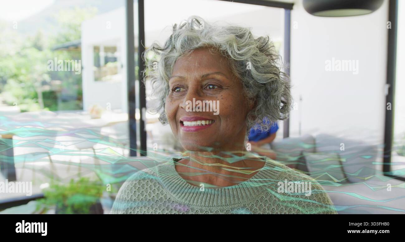 Donna anziana sorridente in maglione in piedi vicino a porte di vetro nel soggiorno, con forme d'onda digitali Foto Stock