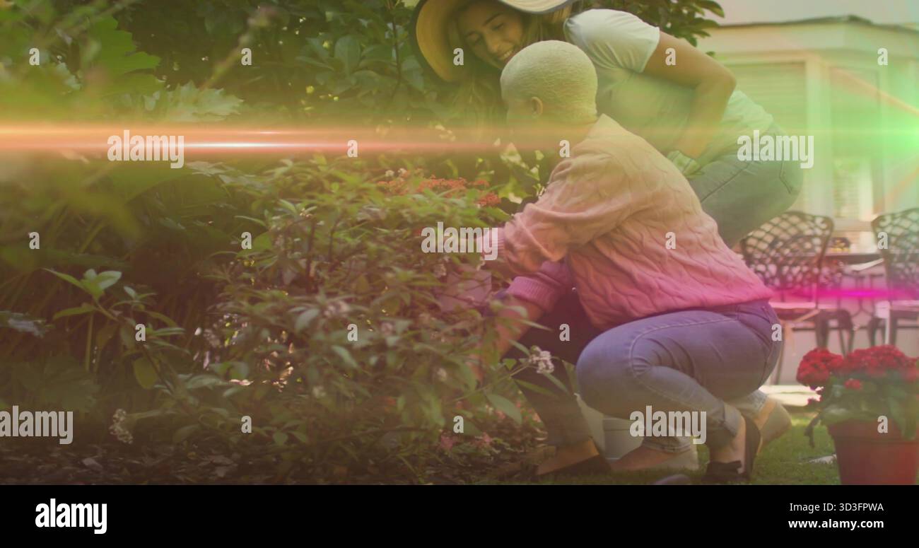 Due donne curano arbusti fioriti nel cortile posteriore, con fiori in vaso rossi, sedie e tavolo da patio Foto Stock