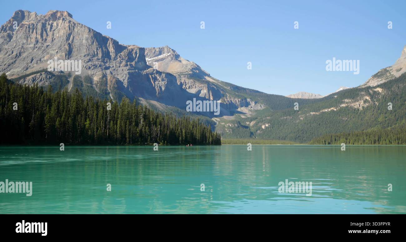 Mostra la scoscesa vetta della montagna che si innalza sopra la linea di alberi sempreverdi sul lago turchese con macchie di neve Foto Stock