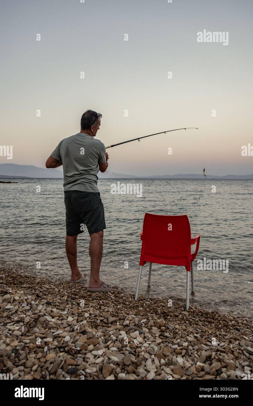 Un uomo si trova sulla costa di ciottoli a pescare con una canna mentre il sole tramonta sul mare sereno. Una sedia rossa nelle vicinanze aggiunge contrasto al tranquillo paesaggio marino Foto Stock