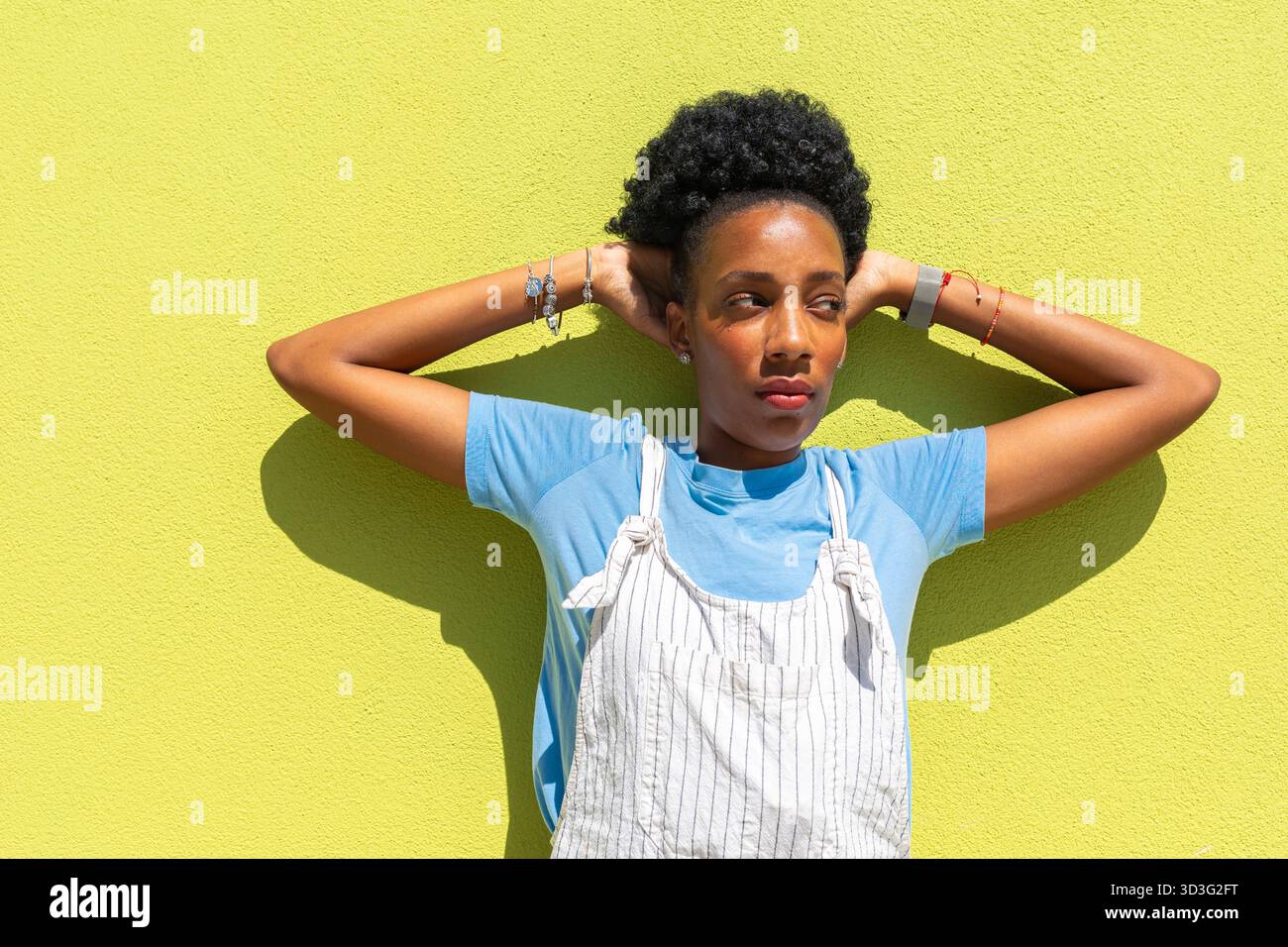 Una donna nera si staglia contro una parete gialla vibrante, indossando una camicia blu e una tuta a righe. Casualmente mette le mani dietro la testa, ammogliandosi Foto Stock