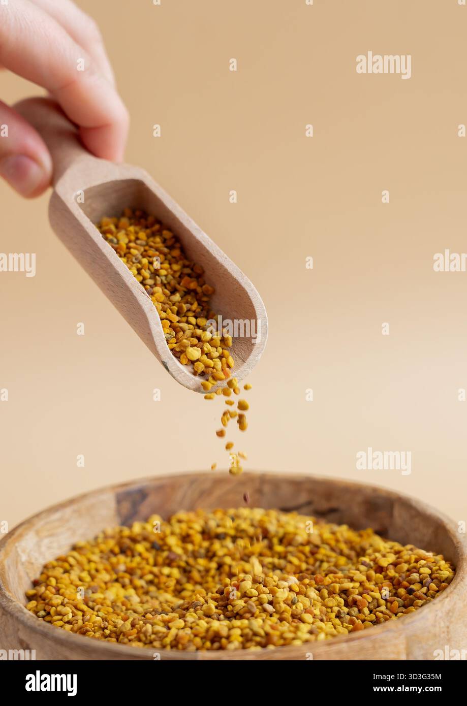 Mano corta e irriconoscibile che regge una paletta di legno di polline d'api naturale su una ciotola di legno su uno sfondo beige liscio. Illustrare i vantaggi di t Foto Stock