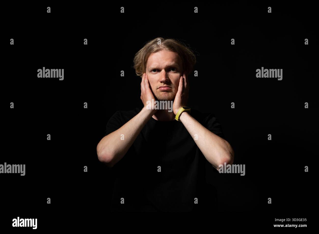 Uomo in camicia nera che tocca il viso con entrambe le mani in un momento di realizzazione sotto una luce drammatica Foto Stock