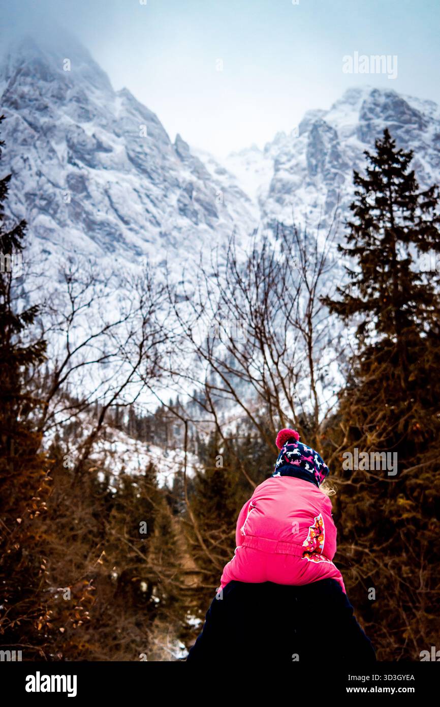 Un bambino seduto sulle spalle del padre. La vista include vette innevate e una foresta sullo sfondo. Foto Stock
