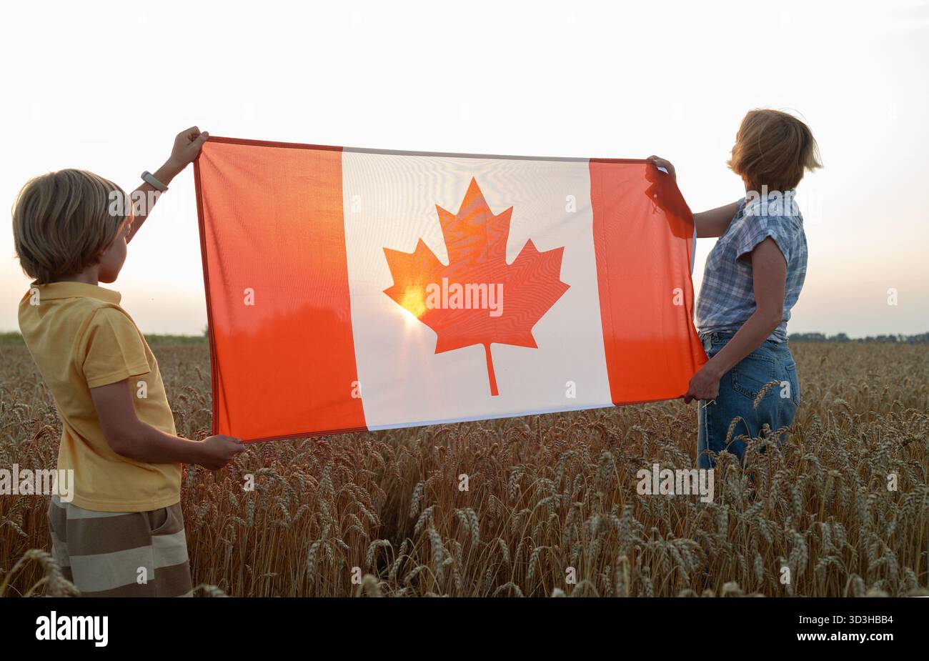 Donna e bambino, madre e figlio, hanno una grande bandiera canadese in piedi in un campo di grano al tramonto. Buon Canada Day Pride, libertà, patriottismo. Travelin Foto Stock