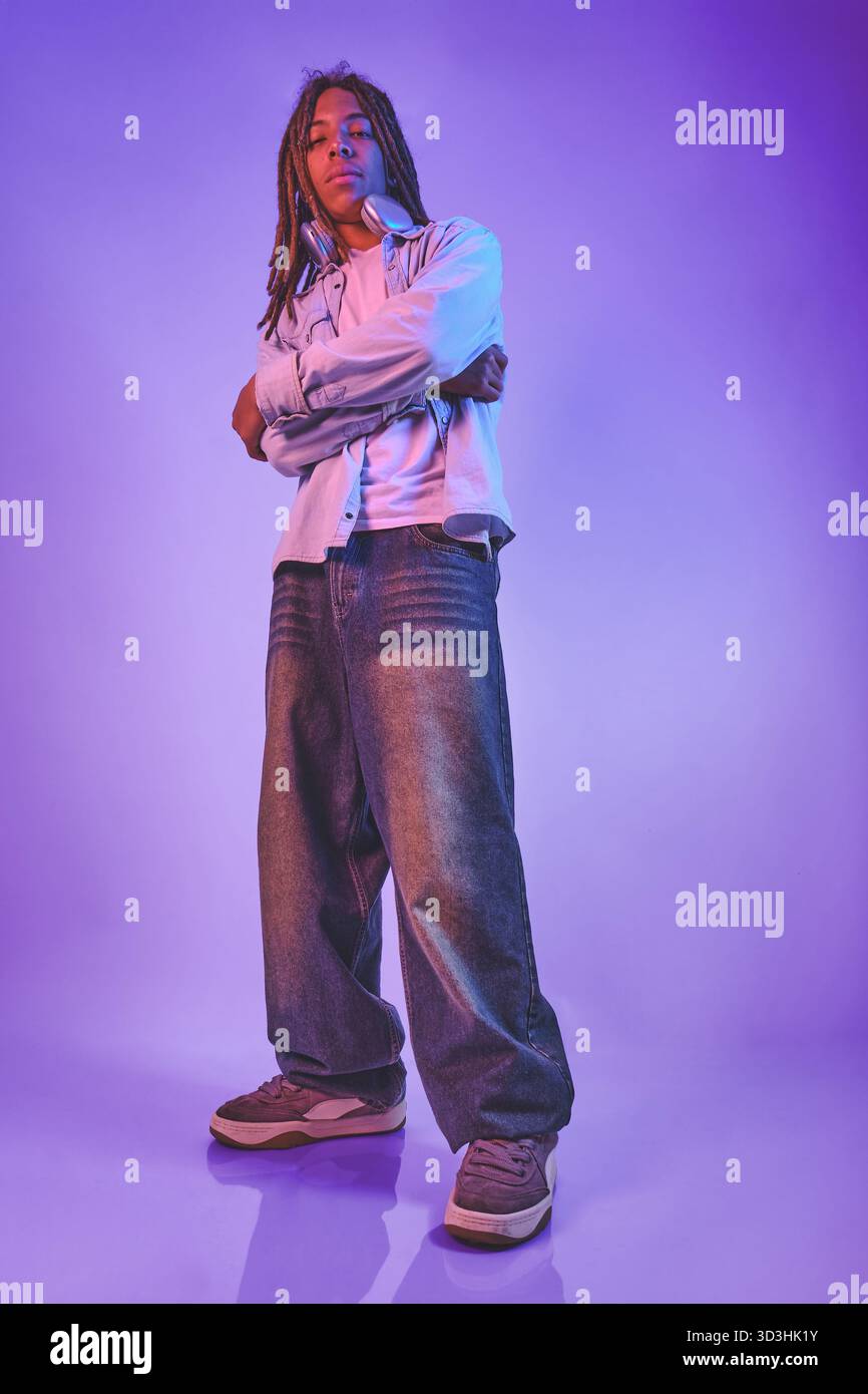 Giovane uomo con dreadlock e cuffie in piedi con sicurezza su uno sfondo viola vibrante Foto Stock