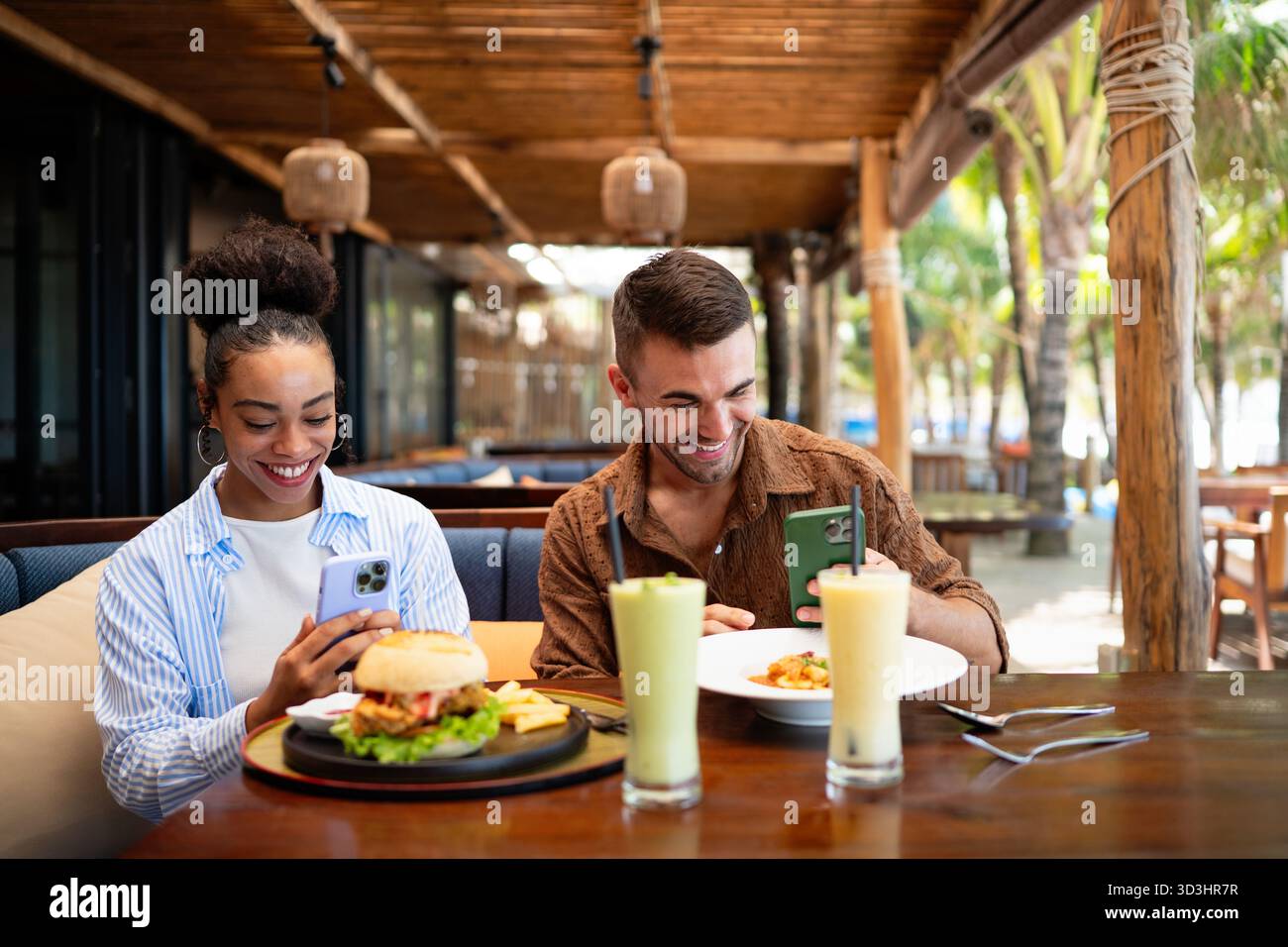 Coppie diverse in un appuntamento gastronomico si godono una cena informale e un momento al telefono, condividendo le vibrazioni soleggiate e le bevande Foto Stock