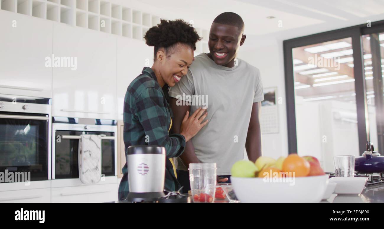 Una coppia che ride si appoggia vicino al bancone della cucina, con frullatore, coltello, fragole e cesto di frutta Foto Stock