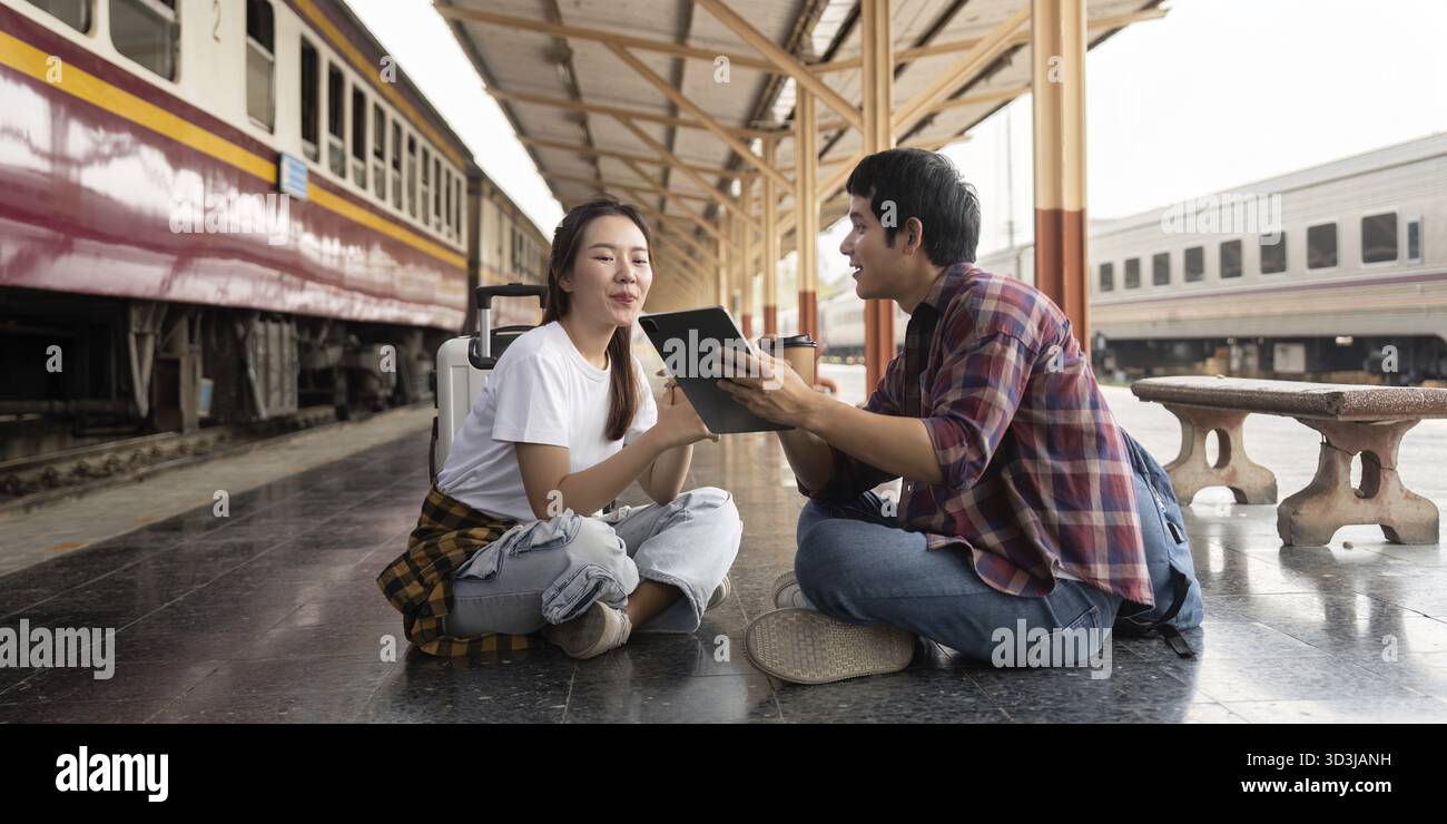 In una vivace stazione ferroviaria, una coppia siede a terra, impegnata in una discussione sui loro prossimi viaggi. L'emozione del loro viaggio in avanti Foto Stock