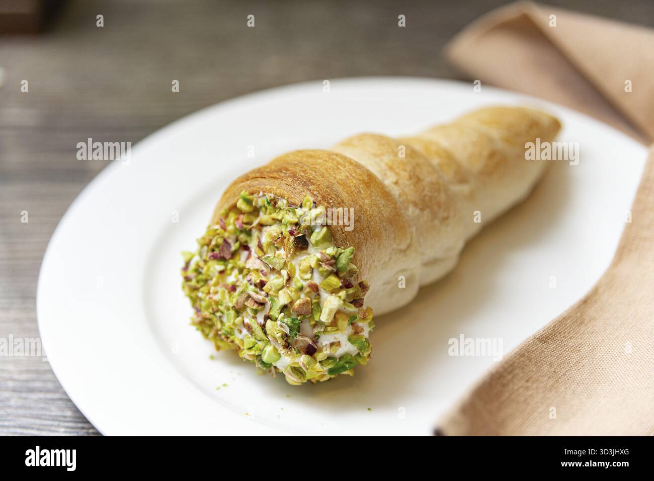 Cibo, Un corno di pasticceria dorato e friabile ripieno di panna dolce e ricoperto di pistacchi schiacciati, elegantemente placcato su un piatto bianco Foto Stock