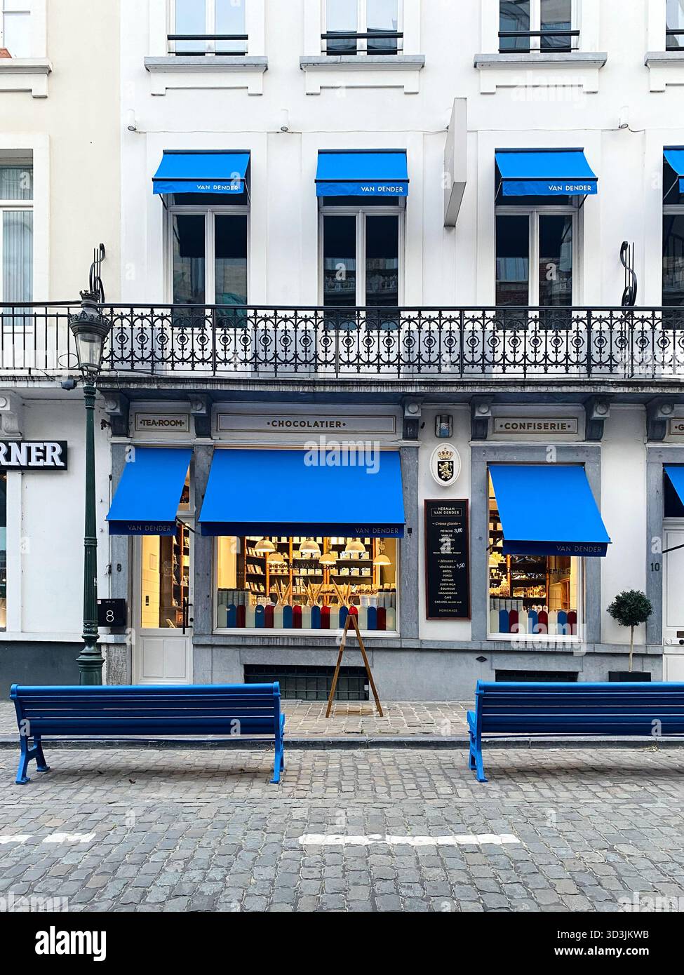 Bruxelles, Belgio - 2 ottobre 2025: Baldacchini blu e bianchi adornano le vetrine di una rinomata pasticceria, completata da panchine blu nelle vicinanze, inviti Foto Stock