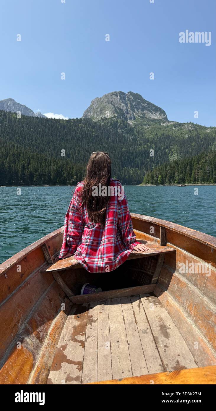 Una donna con una camicia a quadri siede su una barca di legno su un lago montenegrino, di fronte alle montagne e alla foresta mentre si sposta pacificamente sull'acqua. Foto Stock