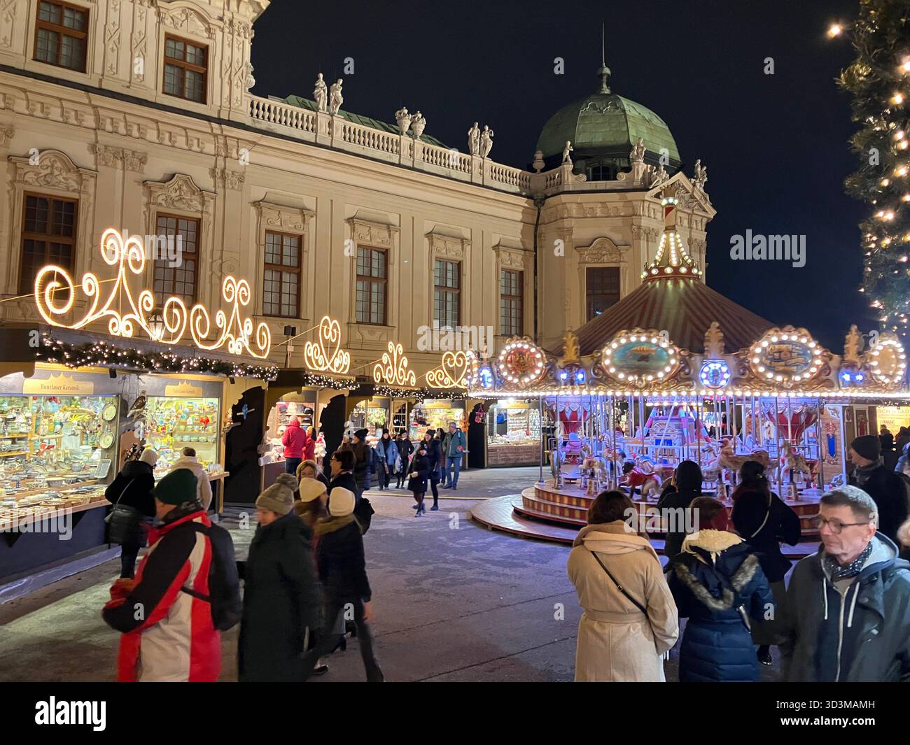 Mercatino di Natale festoso al palazzo Belvedere di Vienna, Austria, con bancarelle illuminate, una giostra e visitatori in una notte d'inverno. - Immagine stock catturata con smartphone