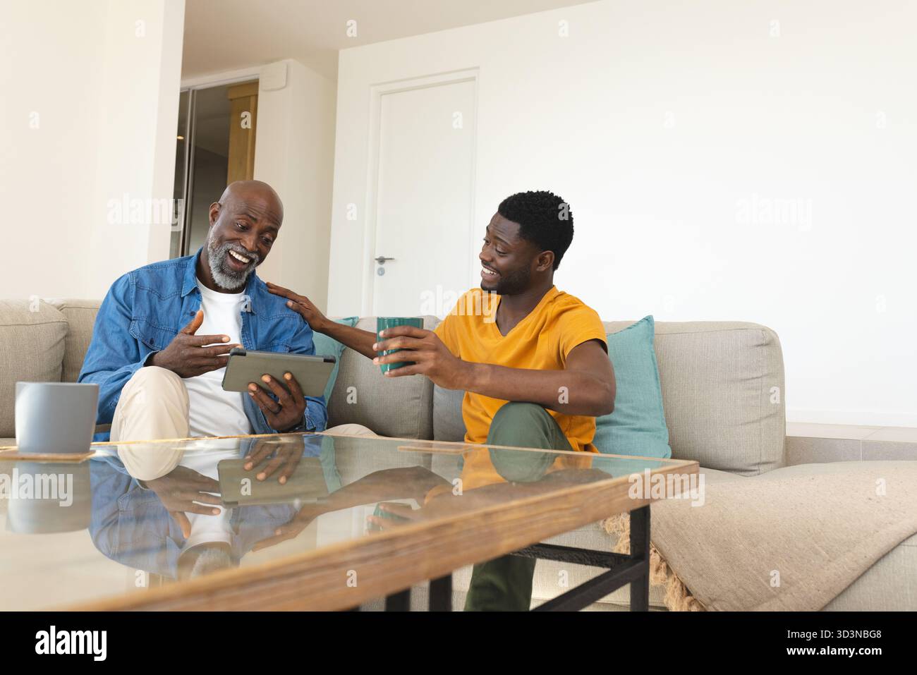 Padre e figlio afroamericani che interagiscono esplorano un tablet in salotto, con tazze in ceramica Foto Stock
