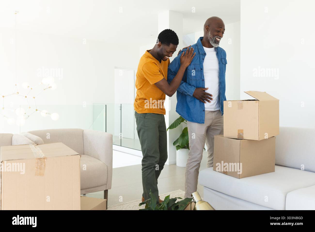 Padre e figlio afroamericani ridendo che disimballano scatole mobili in soggiorno, con una luce appesa Foto Stock
