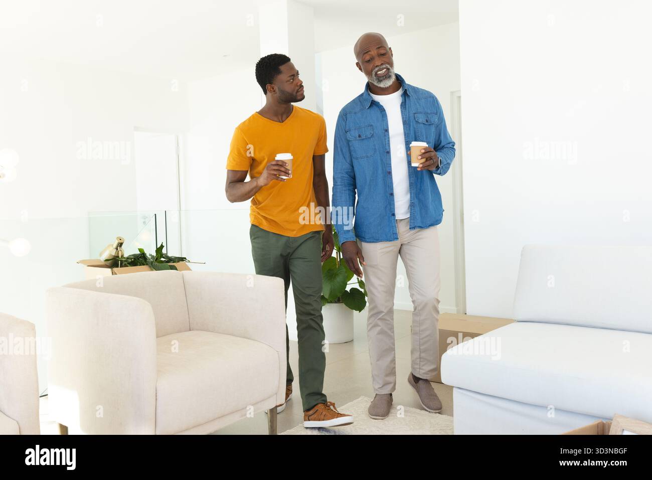 Padre e figlio afroamericani che camminano trasportano tazze da caffè in un luminoso soggiorno, con scatola mobile Foto Stock