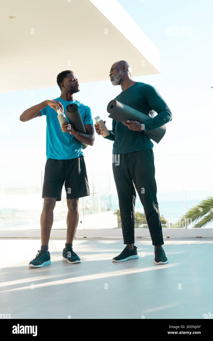 Famiglia afroamericana in piedi con stuoie da yoga e bottiglie d'acqua sulla terrazza affacciata sull'oceano Foto Stock