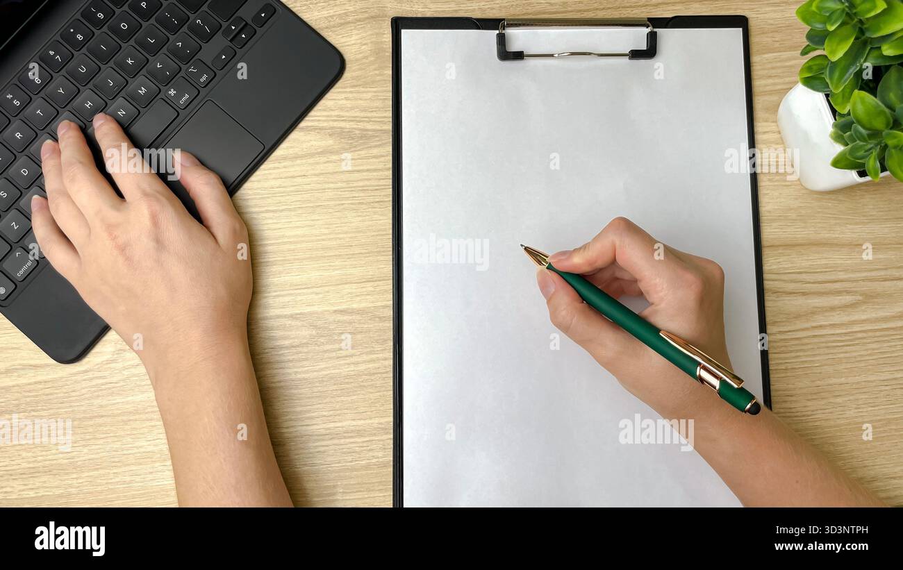 Un uomo che lavora su un computer portatile, digita con una mano e tiene una penna su un foglio di carta bianco con l'altra. - Immagine stock catturata con smartphone
