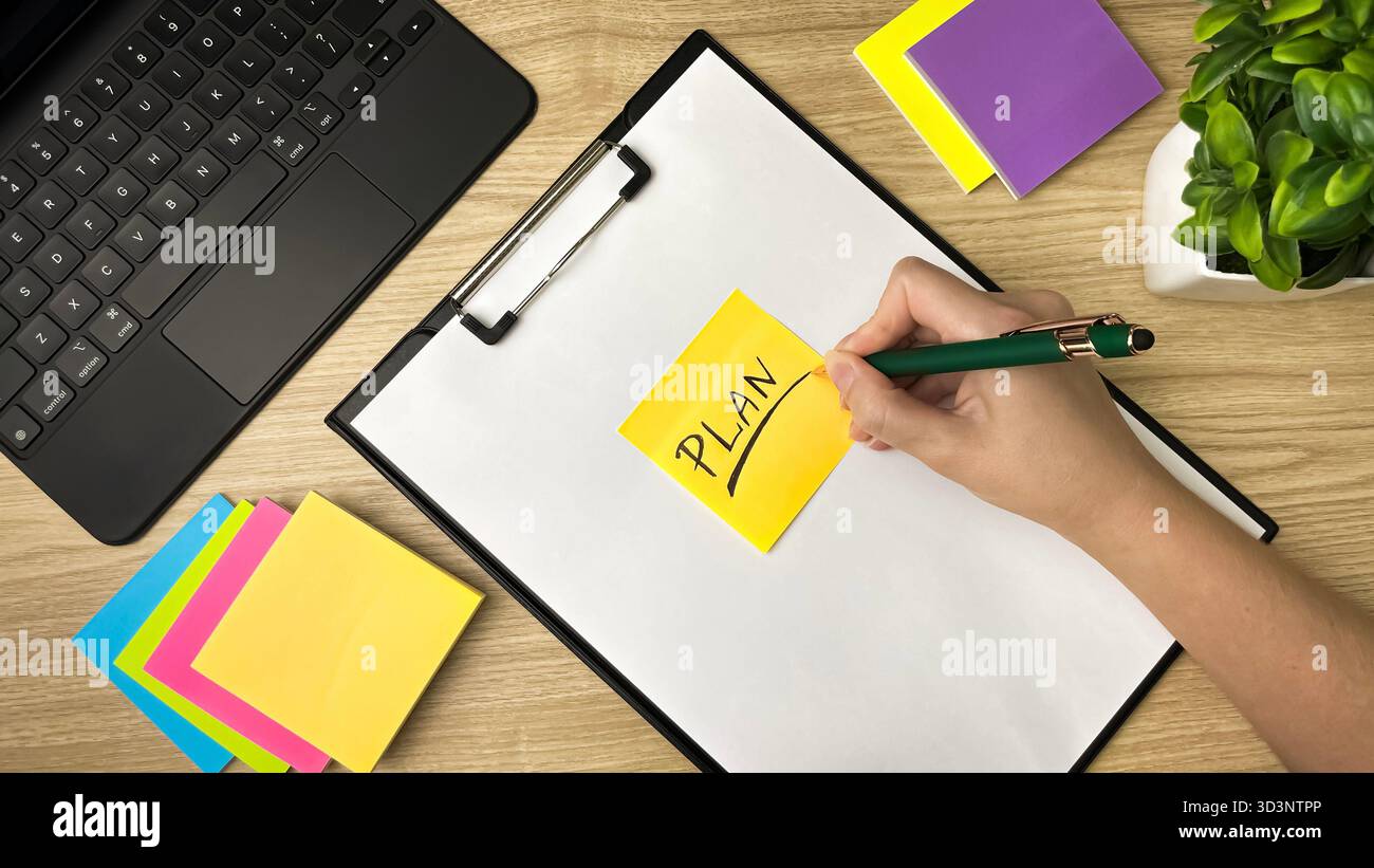 Una scrivania con note colorate, un computer portatile e una mano con una penna. Una nota adesiva recita "piano". Un simbolo di pianificazione, gestione del tempo - Immagine stock catturata con smartphone