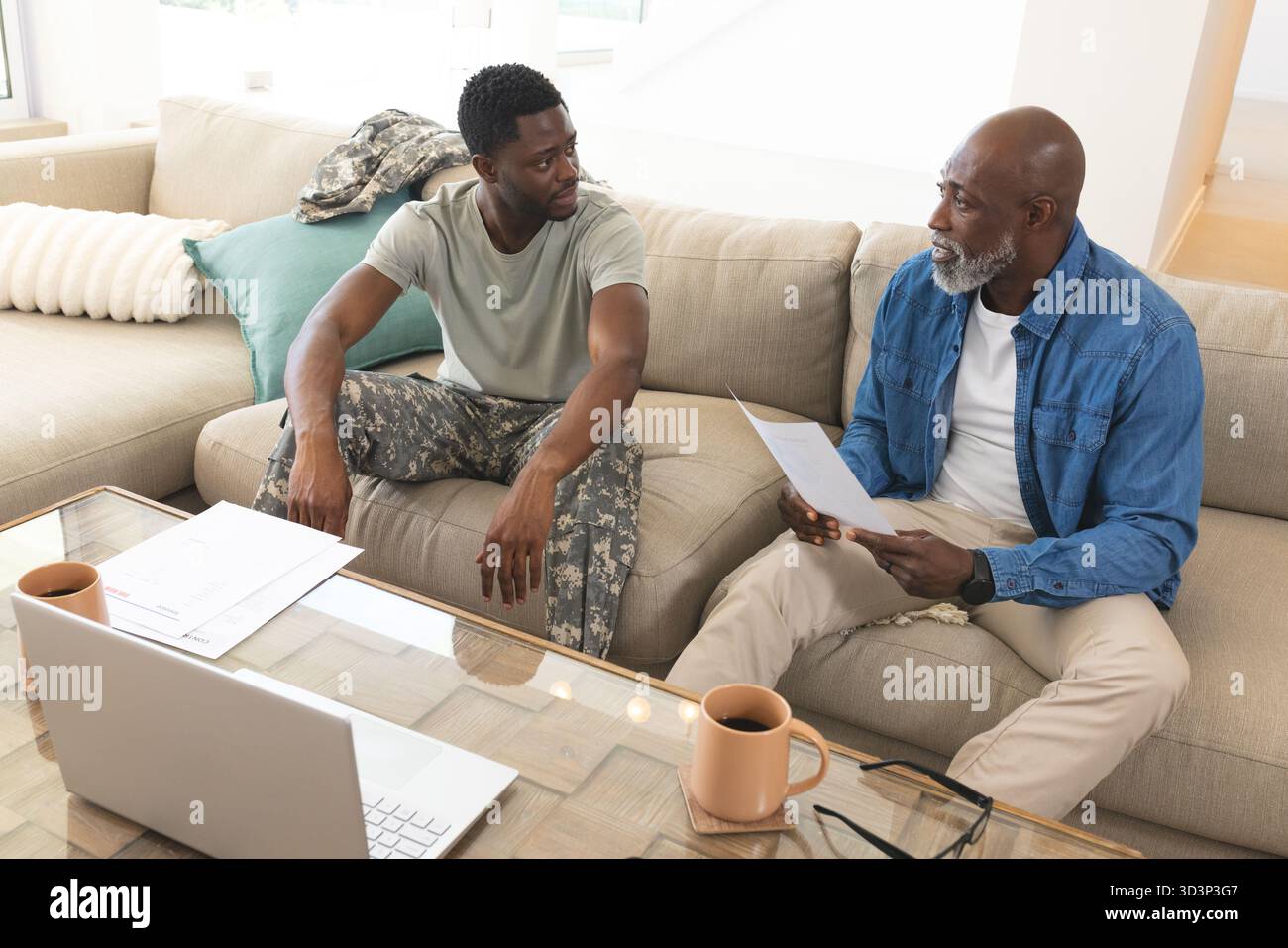 Padre e figlio afroamericani che recensiscono documenti in un luminoso soggiorno, con laptop e tazze Foto Stock