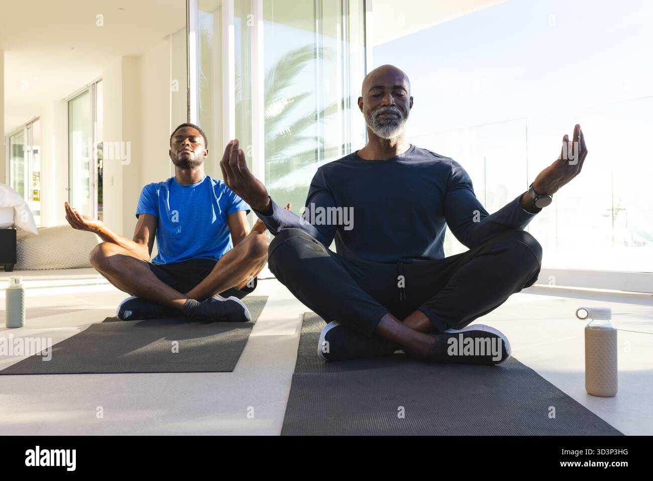 Padre e figlio afroamericani che praticano la meditazione sui tappetini da yoga a casa, con bottiglie d'acqua Foto Stock