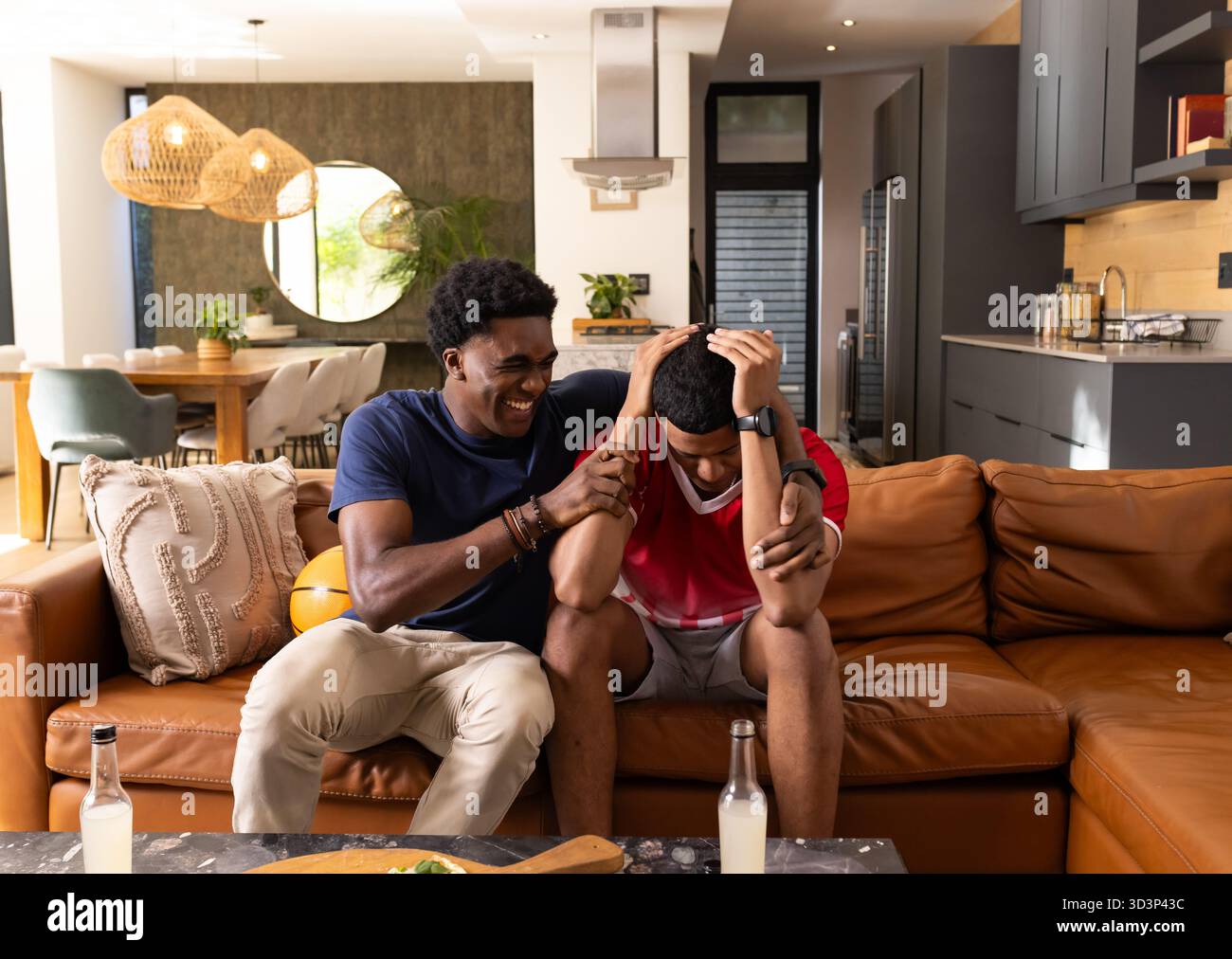 Ridendo diversi amici maschi che si consolano sul divano in una casa a pianta aperta, con calcio e spuntini Foto Stock
