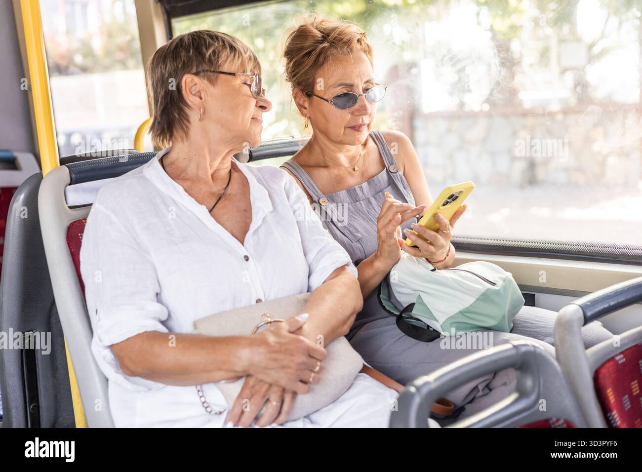 Due amiche mature si siedono insieme su un autobus pubblico, godendosi la compagnia reciproca. Una donna è fidanzata con il suo smartphone mentre l'altra guarda Foto Stock