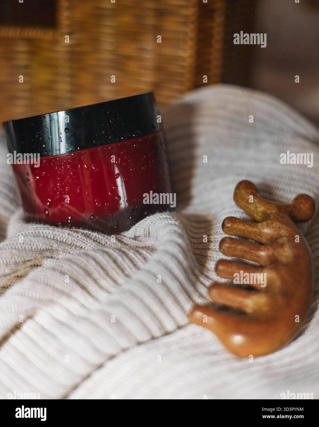 Primo piano di un vaso rosso di prodotto per la cura dei capelli, completato da un pettine di legno, appoggiato su un morbido tessuto beige. L'ambiente evoca calore e lusso Foto Stock
