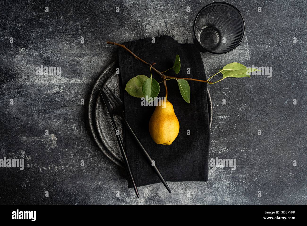 Vista dall'alto di un elegante tavolo con una singola pera gialla su un tovagliolo nero, un piatto in ceramica grigia e utensili neri opachi, il tutto su una superficie testurizzata Foto Stock