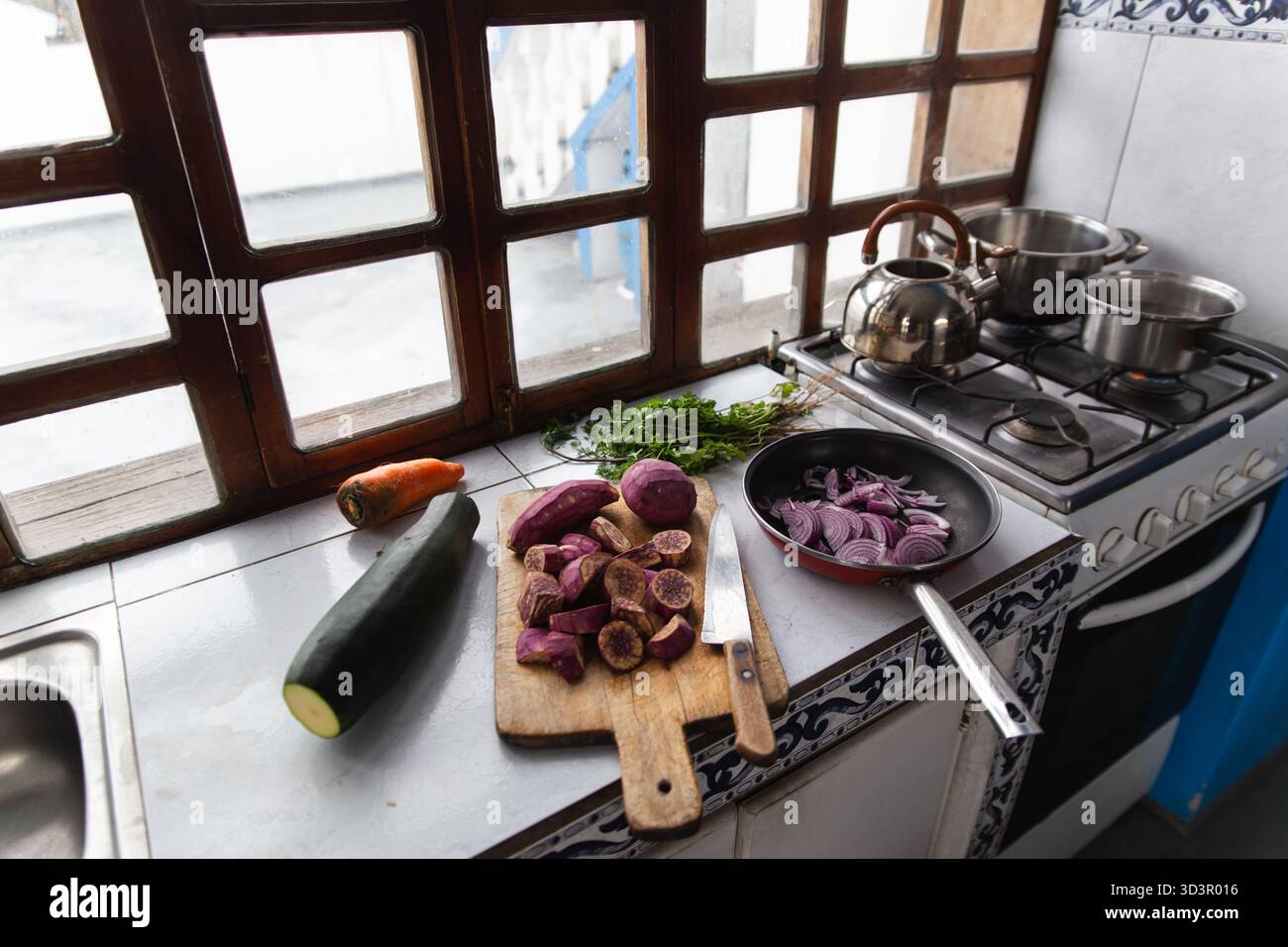 Un'accogliente cucina con verdure fresche come carote, zucchine e patate viola su un bancone. Un tagliere, un coltello e una padella aggiungono t Foto Stock
