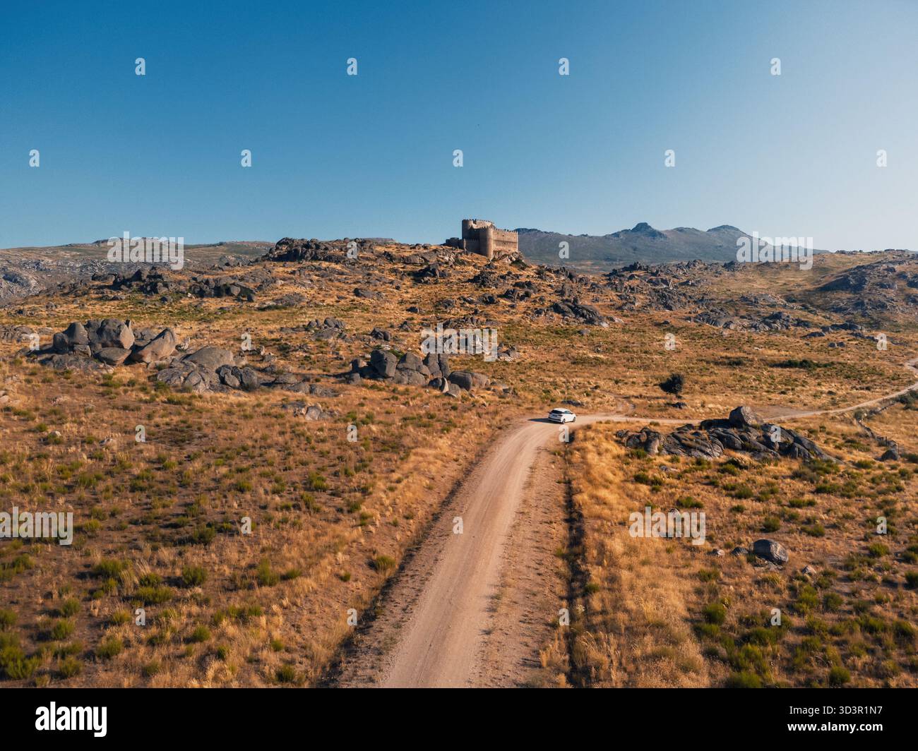 Vista aerea del castello di Manqueospese ad avila, in Spagna, circondato da un paesaggio roccioso e arido. Un'auto bianca percorre una strada polverosa verso le rovine storiche Foto Stock