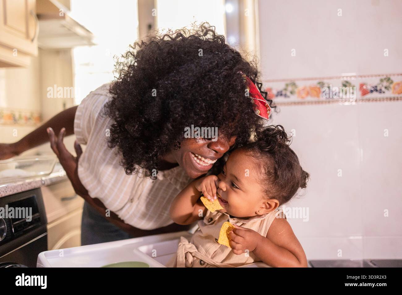 Madre e figlia afroamericane che si godono momenti di qualità a casa in cucina. La madre ride mentre sua figlia stuzzica felicemente su una deliziosa tre Foto Stock
